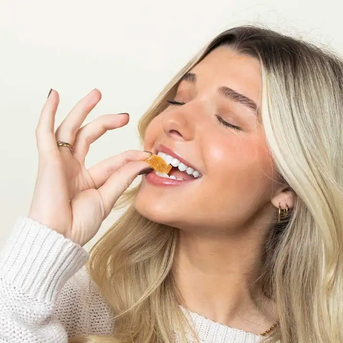 Woman with blonde hair smiling with eyes closed, biting a small orange gummy candy at the moment of a bite.