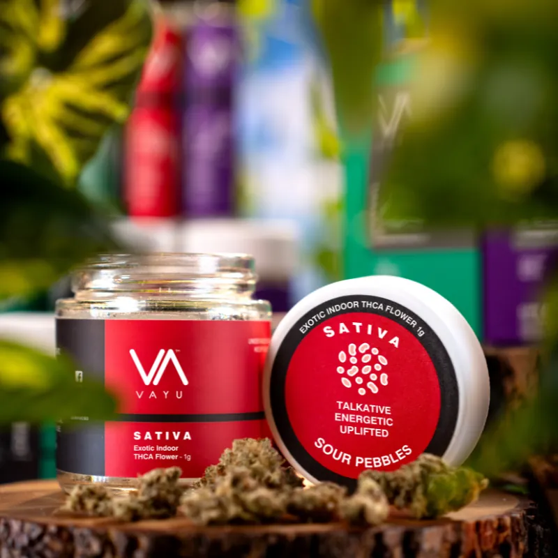 Jar of Vayu Sativa cannabis flower with round red label reading 'Sativa Talkative Energetic Uplifted Sour Pebbles' on a wooden surface.
