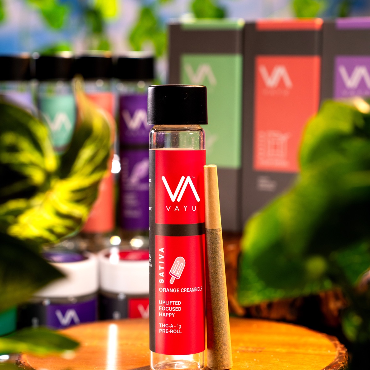Red Vayu Sativa vape bottle on a wooden surface with a pre-roll beside it; blurred colorful packaging in the background.